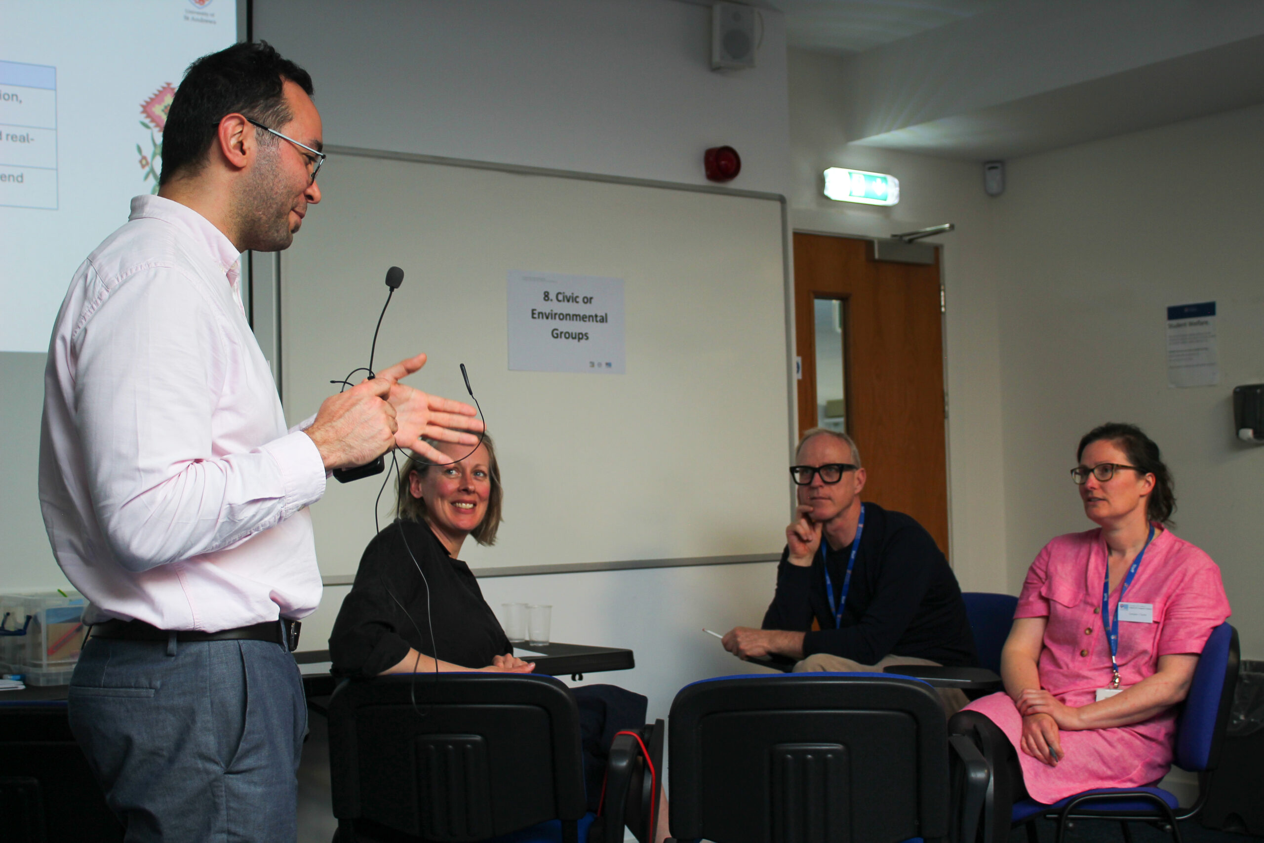 the presenter holding the microphone and speaking while 3 participants in seats look and listen to the speaker